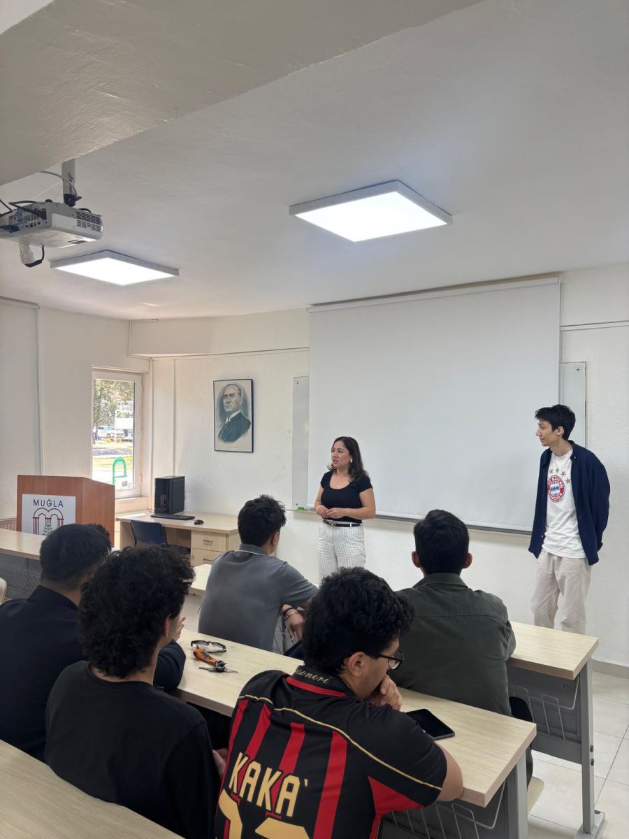 The photo, taken in a classroom, shows two people standing at the front and students sitting at their desks. The students are facing the front of the room. The classroom contains a projection screen, a ceiling-mounted projector, a window, a lectern, and a framed portrait on the wall.